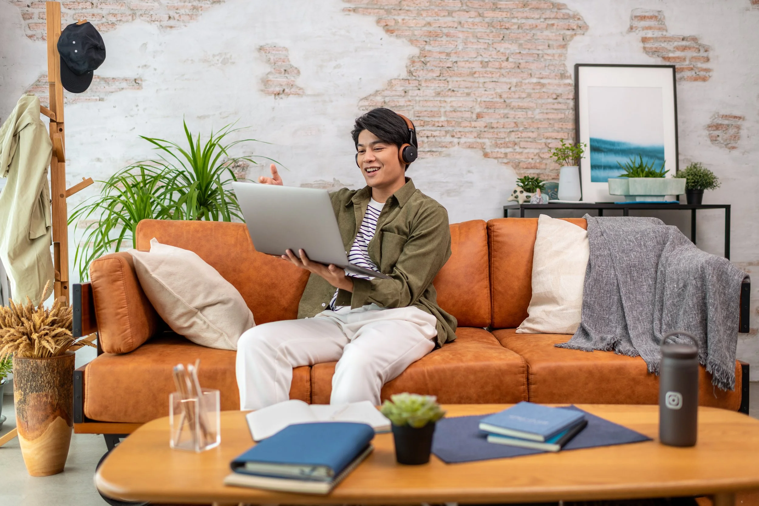 A young man with dark hair, wearing headphones, a striped shirt, and an olive jacket, sitting on an orange sofa in a modern living room, smiling and looking at a laptop.