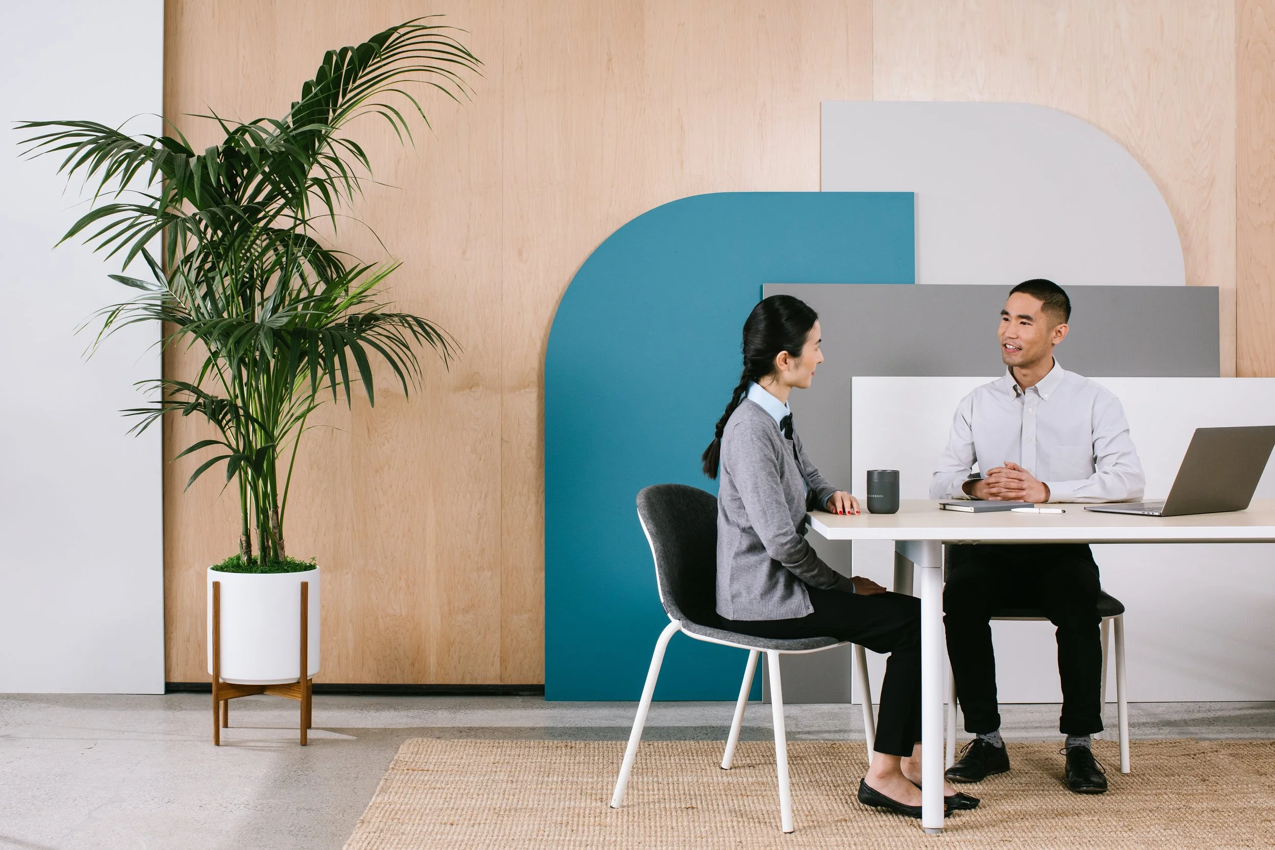 Two people sitting across from each other at a white table in an office or meeting space, engaged in conversation. A large potted plant is on the left, and geometric wall art decorates the background.
