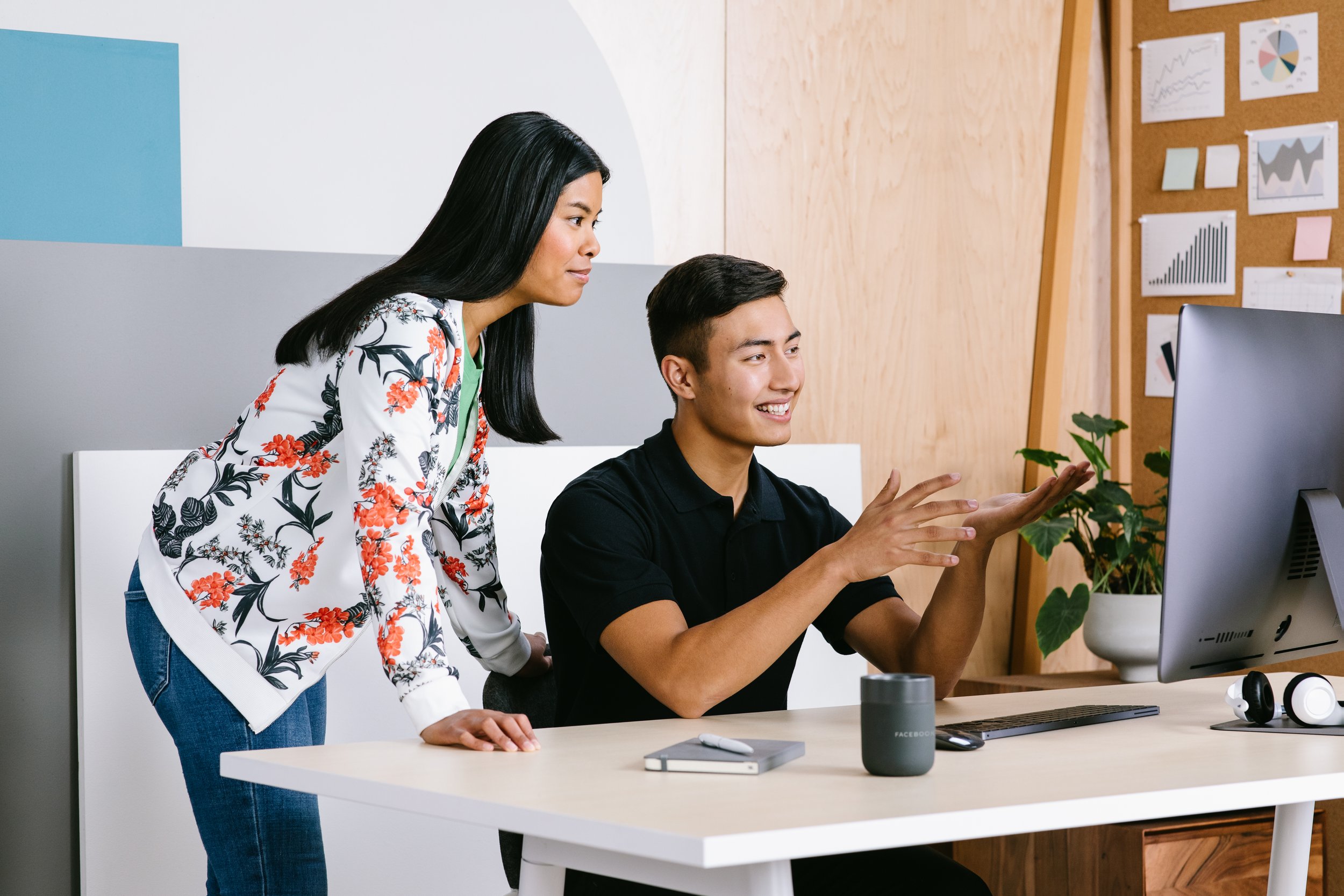 A woman standing next to a seated man shows him something on a computer screen in an office.