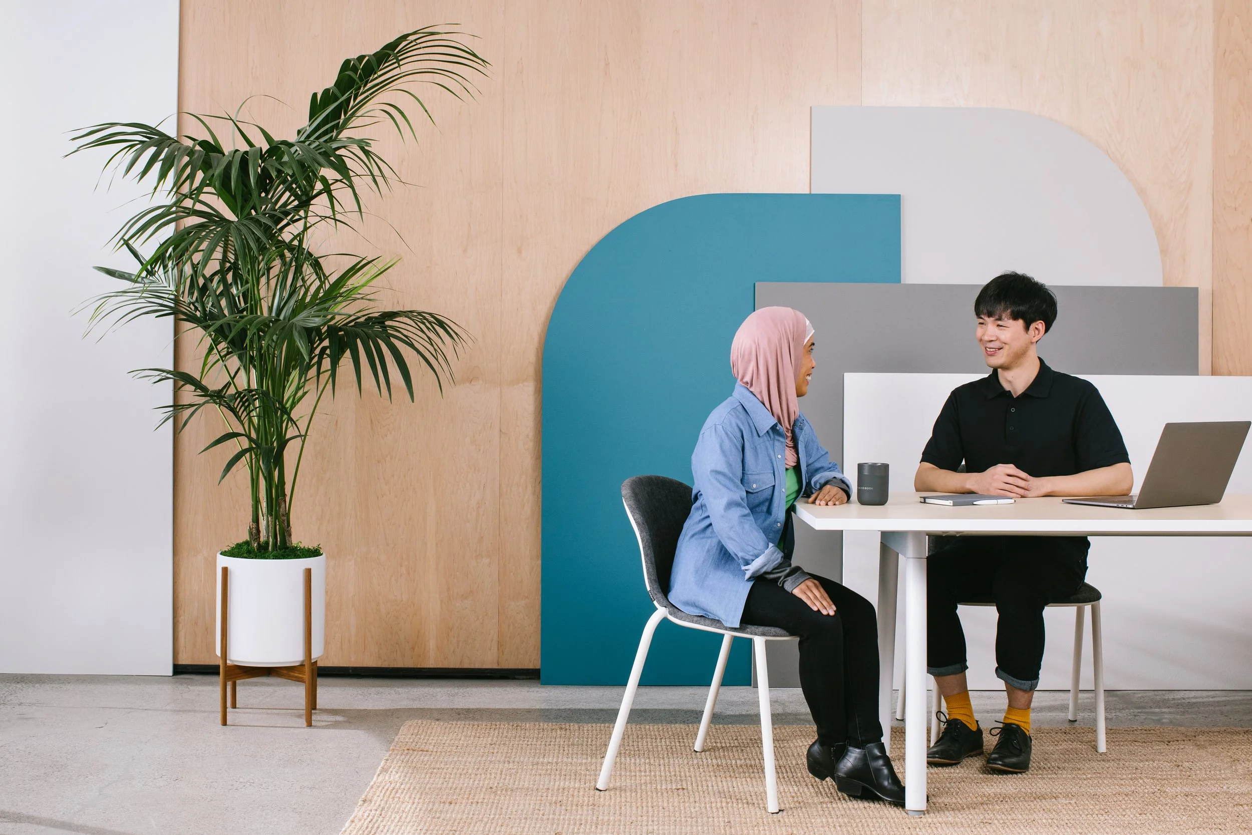 Two people sitting and talking at a white table in a modern office or meeting space, with a large potted plant on the left and colorful geometric wall art behind them.