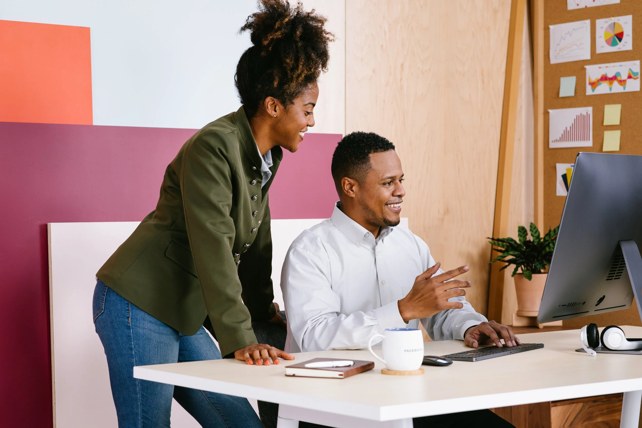 A woman leaning over a desk and smiling while talking to an excited man seated in front of a computer. The desk has a coffee mug, notebook, and headphones. The background features charts and graphs on a corkboard.