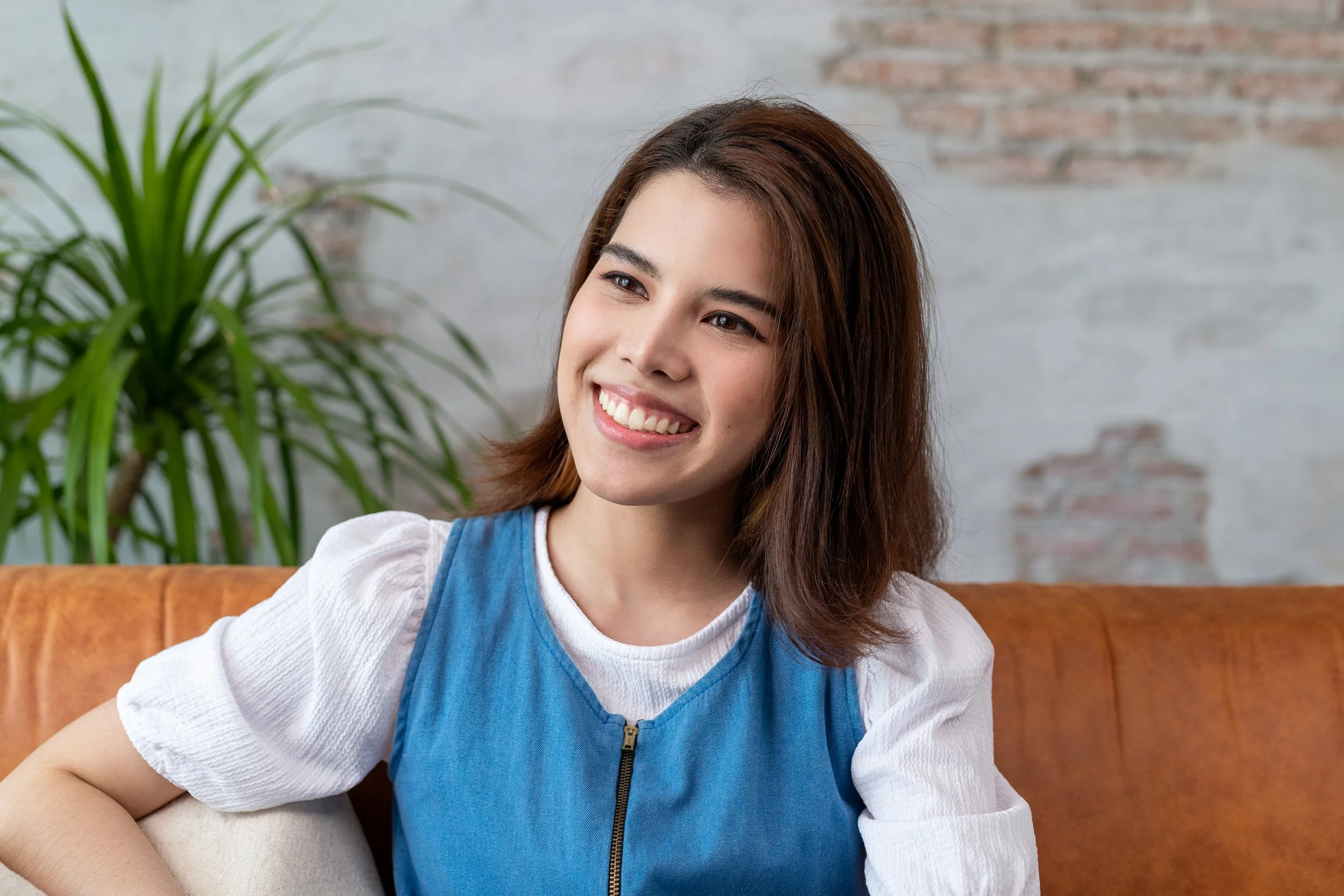 A young woman with brown hair smiling and wearing a blue zip-up top over a white shirt, sitting on a brown leather couch in a room with a brick wall and a large green plant.