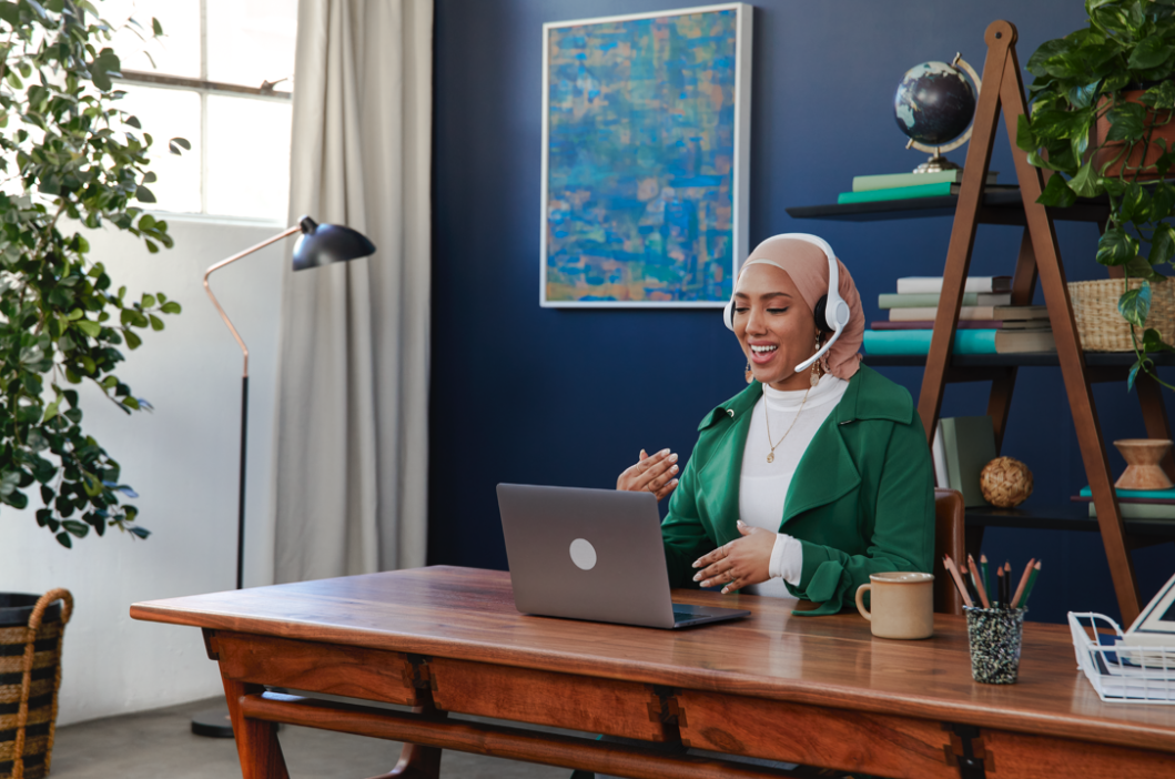 A woman wearing a hijab, headphones, and a green jacket sitting at a wooden desk with a laptop, smiling and speaking during a video call in a bright room with a blue wall, a potted plant, a bookshelf, and artwork.