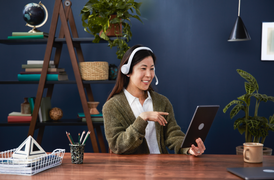 A woman wearing headphones and a white shirt with a green cardigan sitting at a wooden desk, holding a tablet and smiling, in a home or office setting with plants and books in the background.