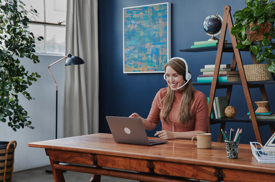 Woman wearing a headset smiling while working on a laptop at a wooden desk in a home office with a blue accent wall and shelves with books and decor.