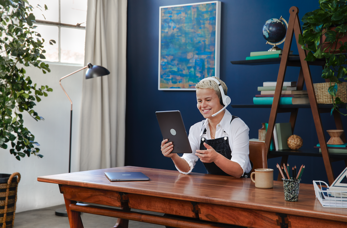 A woman with short blonde hair wearing a headset and black apron, sitting at a wooden desk, smiling while looking at a tablet.