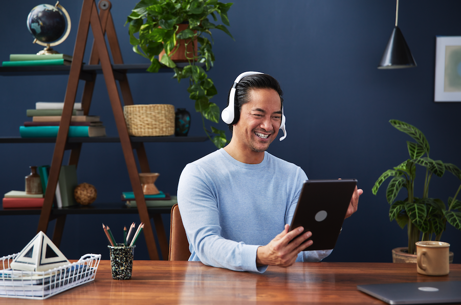 A man wearing a light blue sweater and a headset, smiling and looking at a tablet, sitting at a wooden desk in a home office with plants, shelves, and decor.