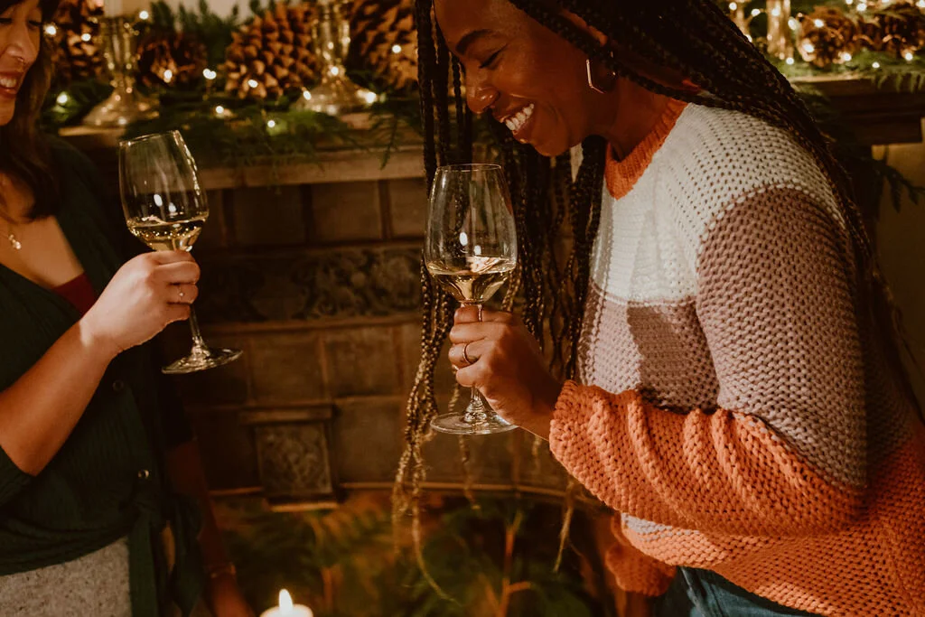 Two women holding wine glasses and smiling at each other at a festive event with holiday decorations.