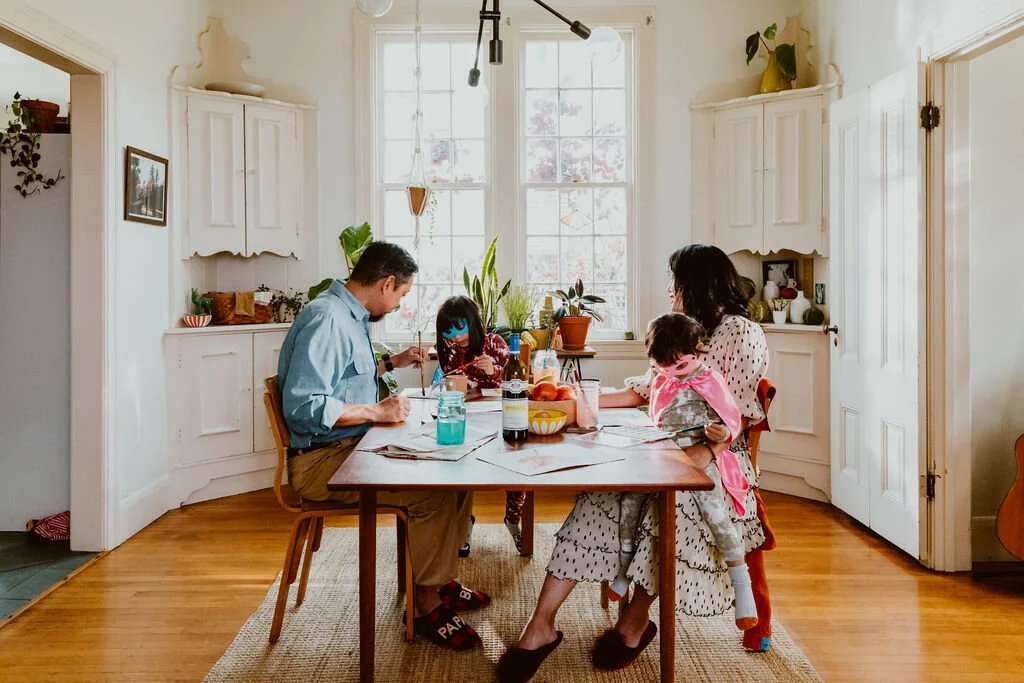 Family of four sitting at a wooden dining table in a bright kitchen, with two children and two adults, enjoying a meal together.