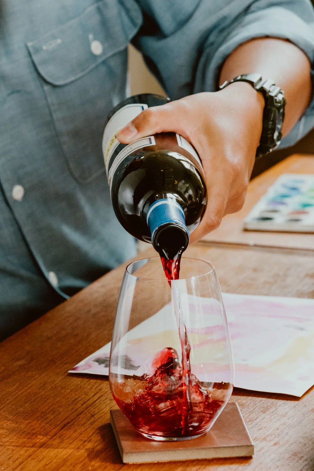 Person pouring red wine into a clear glass on a wooden table.