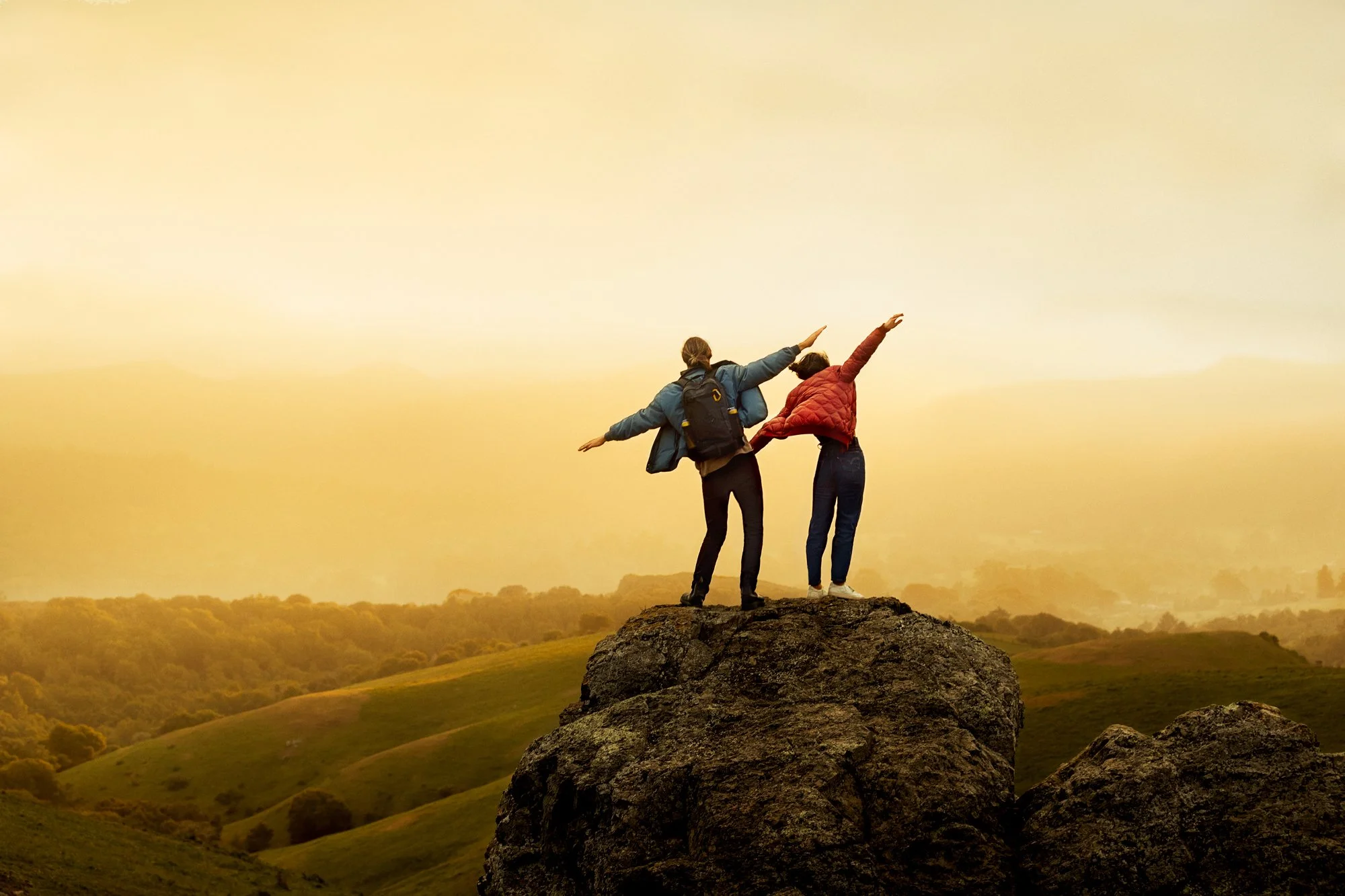 Two women standing on a large rock at sunset, one wearing a blue jacket and backpack, the other in a red quilted jacket, raising their arms in excitement against a yellow-orange sky and rolling green hills.
