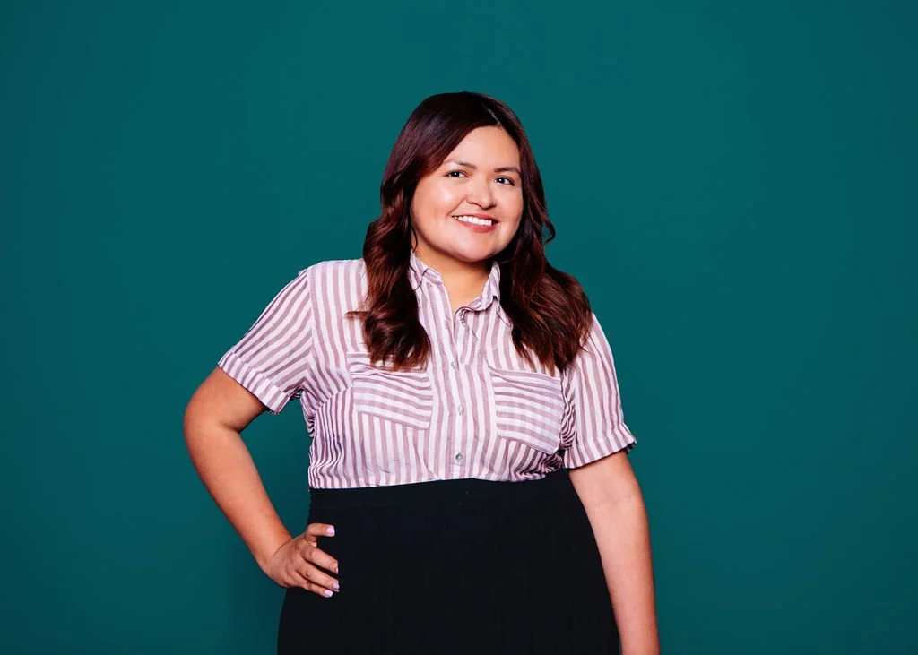 A smiling woman with long, wavy brown hair, wearing a pink and white striped short-sleeve button-up shirt and black pants, standing with one hand on her hip against a teal background.