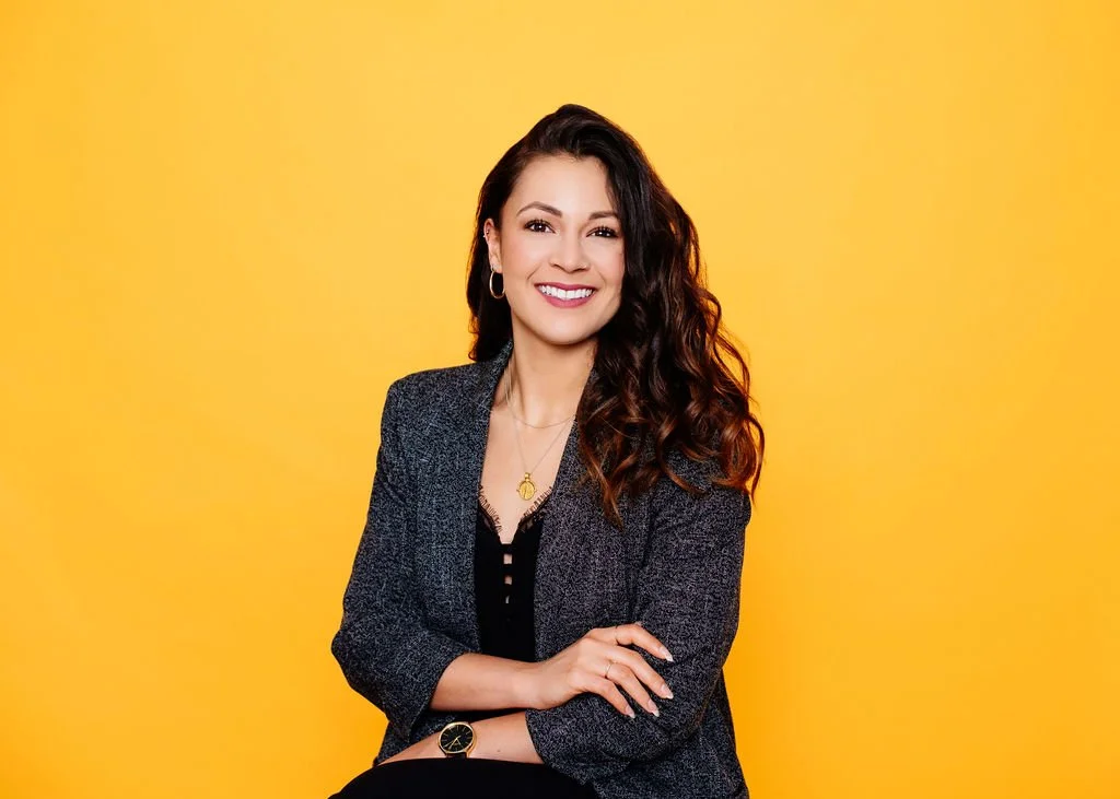 A woman with long curly hair, wearing a gray blazer over a black top, sitting with crossed arms against a yellow background, smiling.