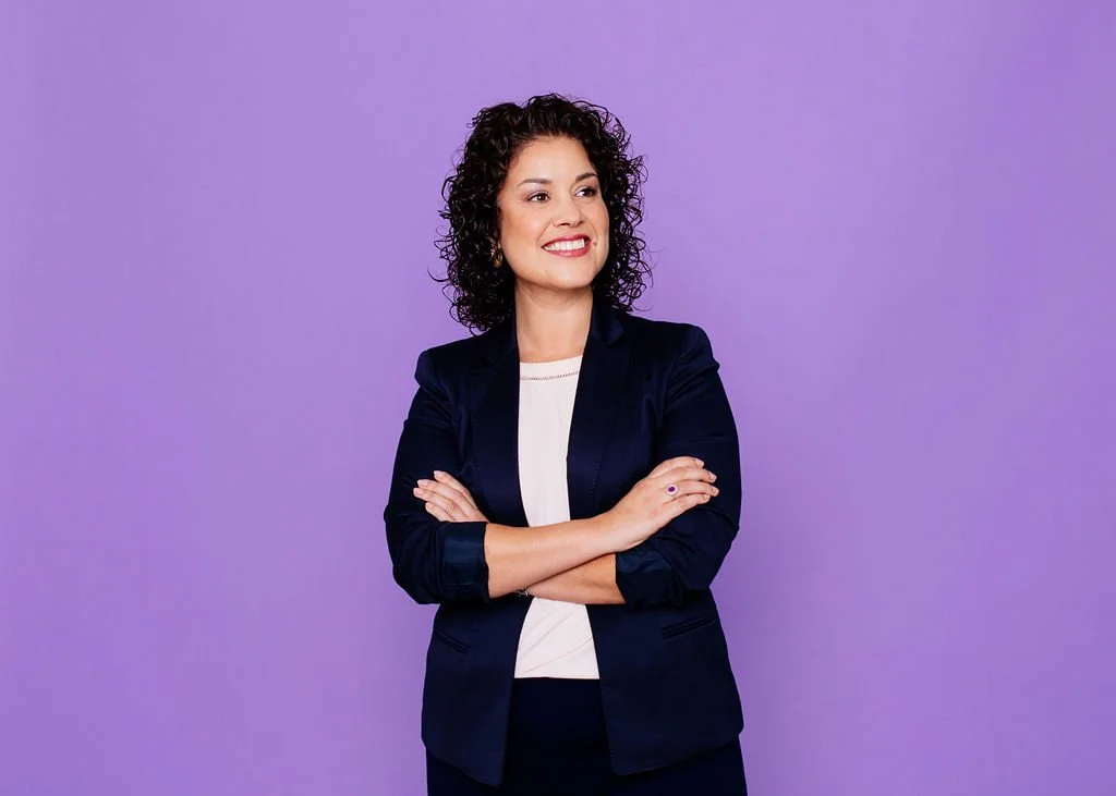 A woman with curly brown hair wearing a black blazer and white top, standing with her arms crossed in front of a purple background, smiling confidently.
