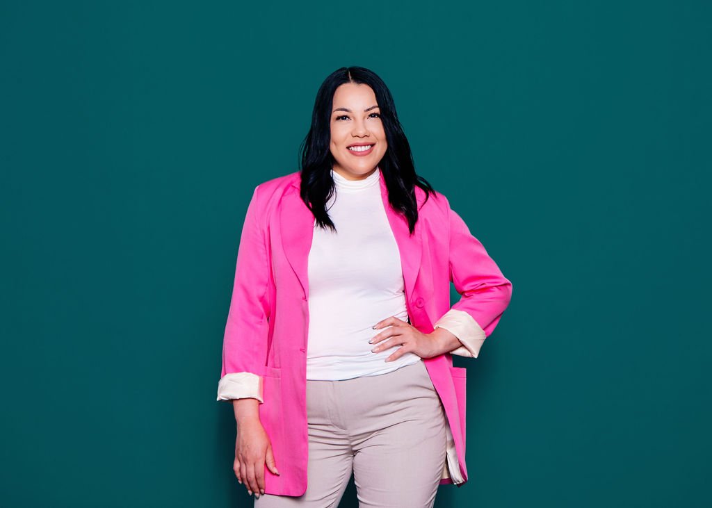 A woman with black hair smiling, wearing a bright pink blazer over a white top and beige pants, standing against a solid green background.
