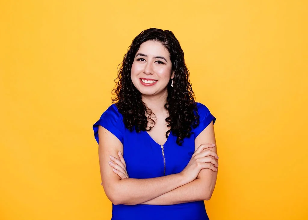 A woman with curly dark hair wearing a royal blue dress, smiling with crossed arms against a yellow background.
