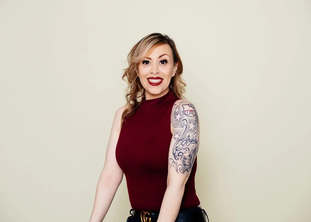 A woman with wavy blonde hair, red lipstick, wearing a sleeveless burgundy top, and has tattoos on her right arm, standing against a light beige background, smiling at the camera.