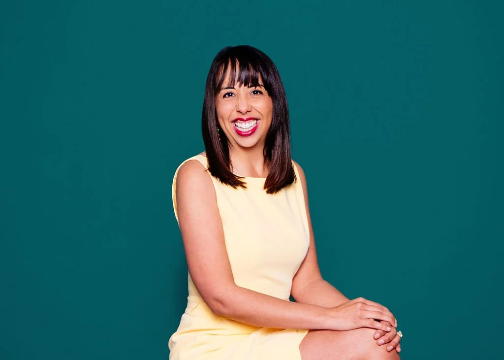 A woman with dark brown hair and bangs, wearing a sleeveless yellow dress, smiling and sitting with her hands on her lap against a teal background.