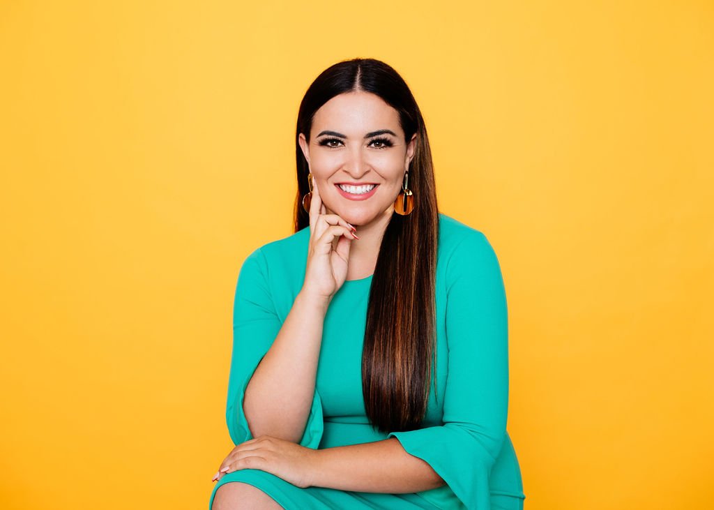 Portrait of a smiling woman with long brown hair wearing a teal dress and gold earrings, sitting against a yellow background.