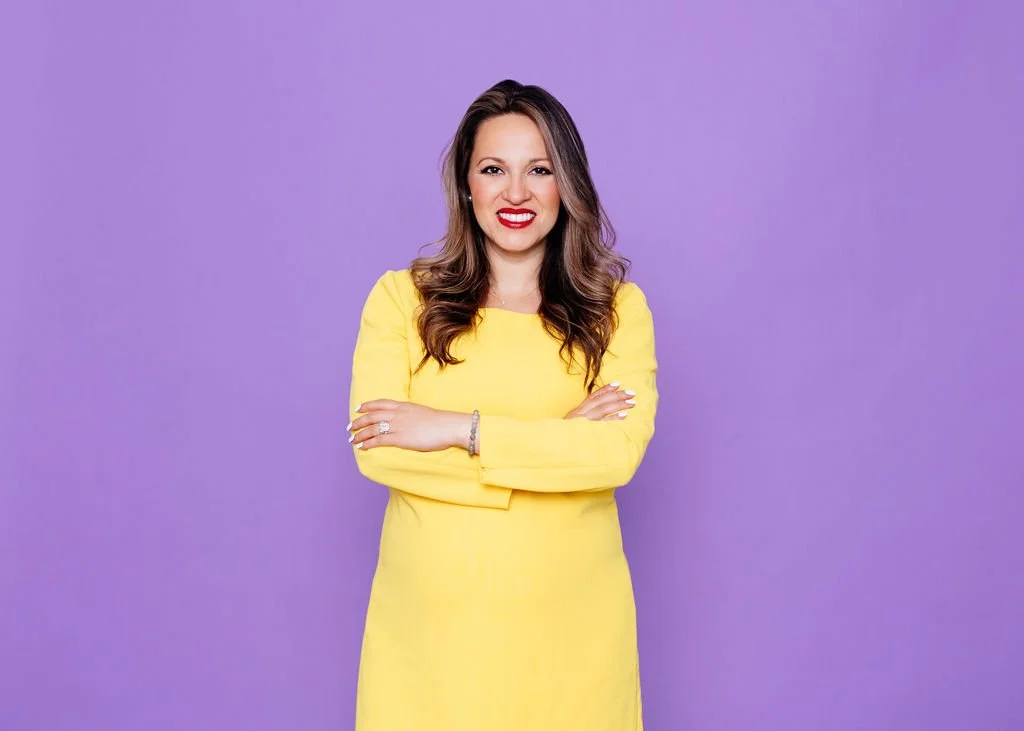 A woman with long wavy brown hair wearing a bright yellow dress, smiling with her arms crossed, standing against a purple background.