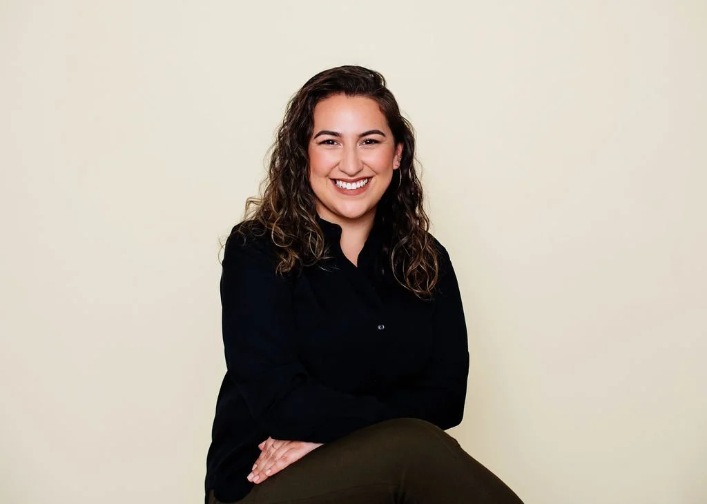 A woman with curly brown hair, wearing a black shirt, smiling and sitting against a plain light-colored background.