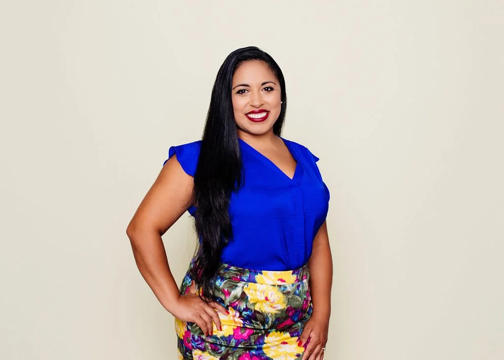 A woman with long dark hair, smiling, wearing a blue top and a colorful floral skirt, standing against a plain light-colored background.
