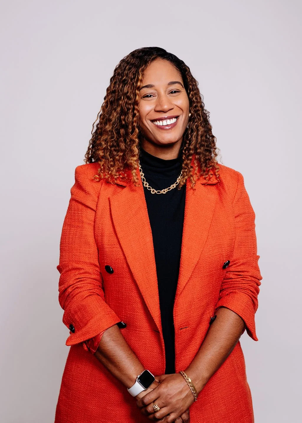 A woman with curly hair wearing an orange blazer and a black top, smiling, standing against a plain gray background.