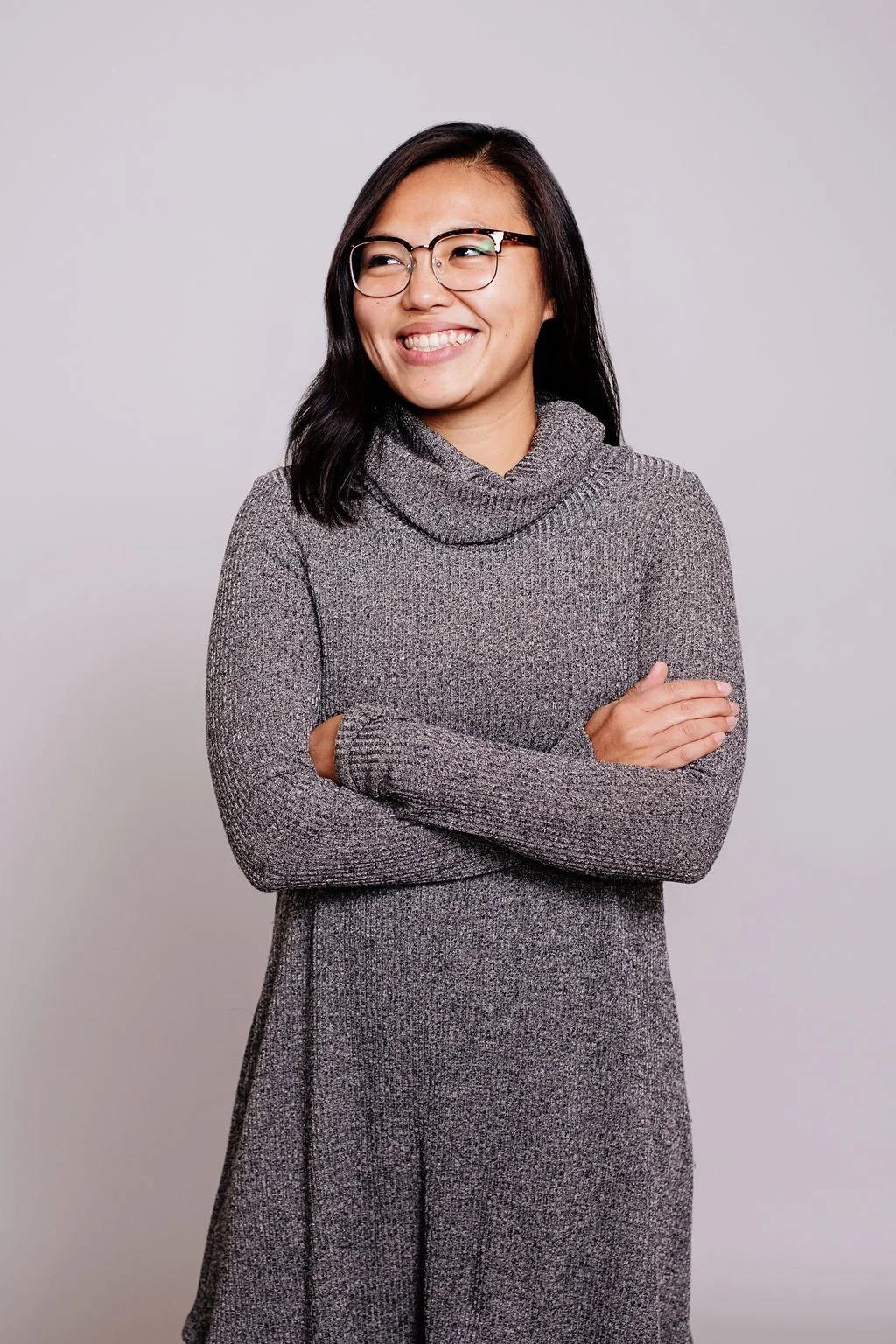 A woman with shoulder-length black hair, glasses, and a grey sweater with a turtleneck, smiling and crossing her arms, standing against a plain light grey background.