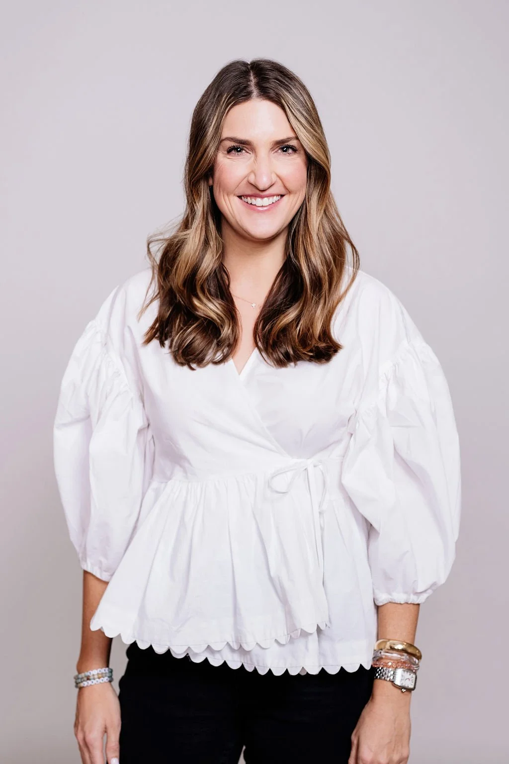 A woman with light brown hair wavy hairstyle, wearing a white blouse with puffed sleeves and black pants, smiling against a plain gray background.