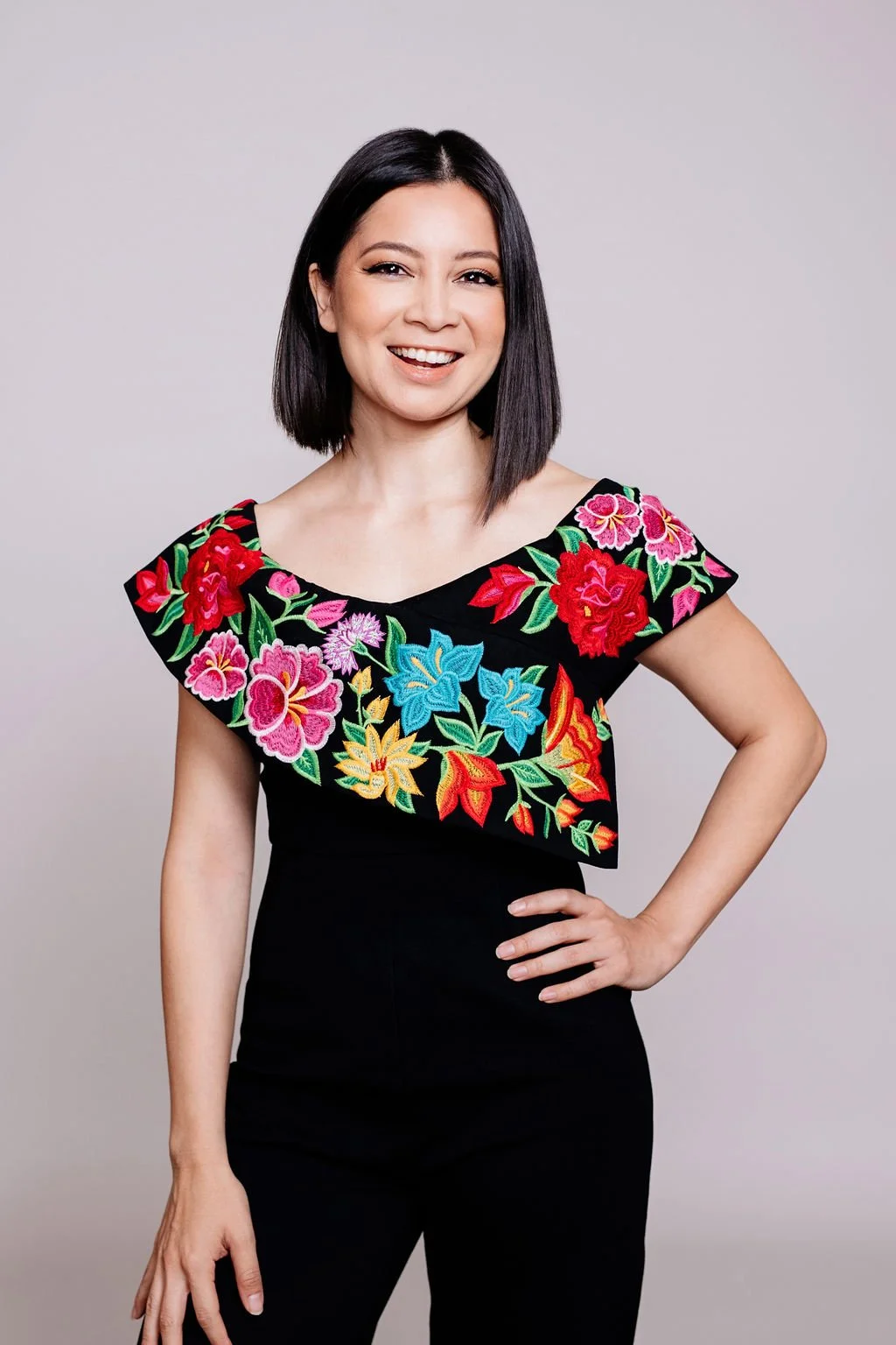 Woman with shoulder-length black hair, smiling, wearing a black top with colorful embroidered floral patterns on the shoulders and upper chest, standing against a light gray background.