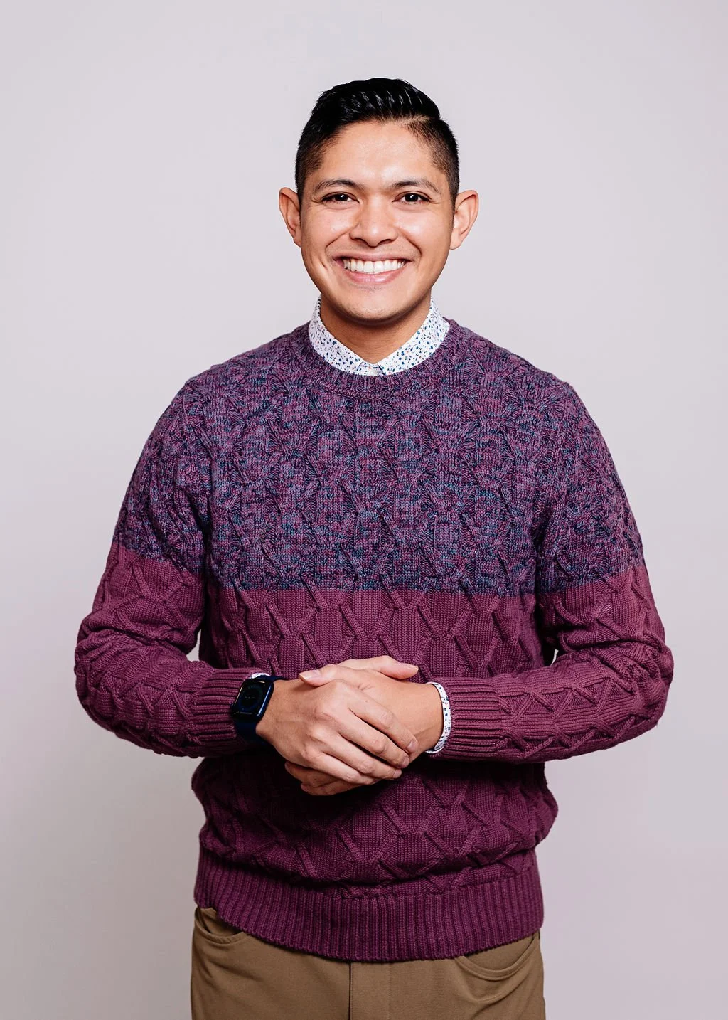 A smiling young man with short dark hair, wearing a purple and pink cable-knit sweater over a collared shirt, and tan pants, standing against a plain light gray background.