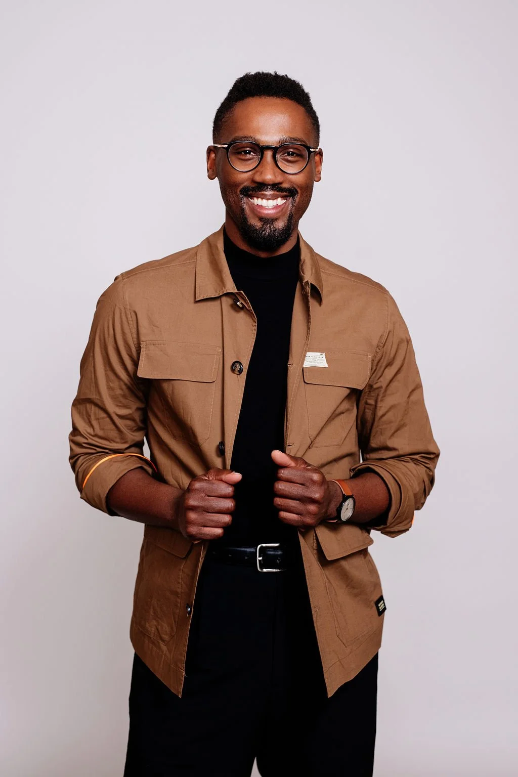 A smiling man with glasses wearing a brown jacket over a black turtleneck, standing against a plain light gray background.