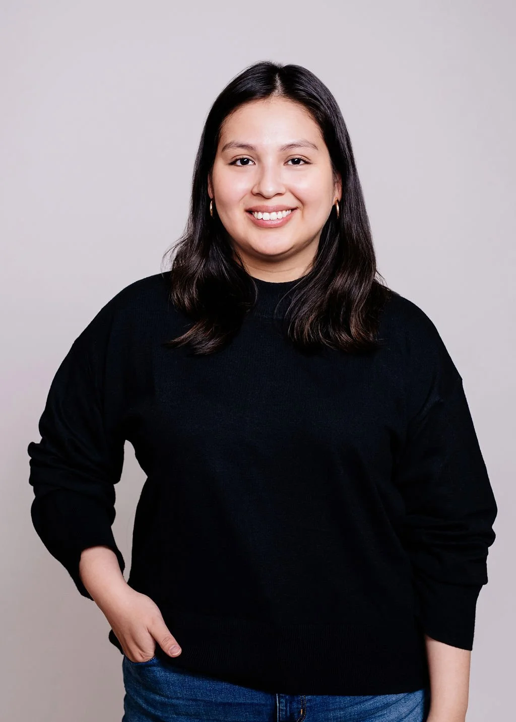 A young woman with shoulder-length dark hair, smiling, wearing a black sweater and blue jeans, standing against a plain light gray background.