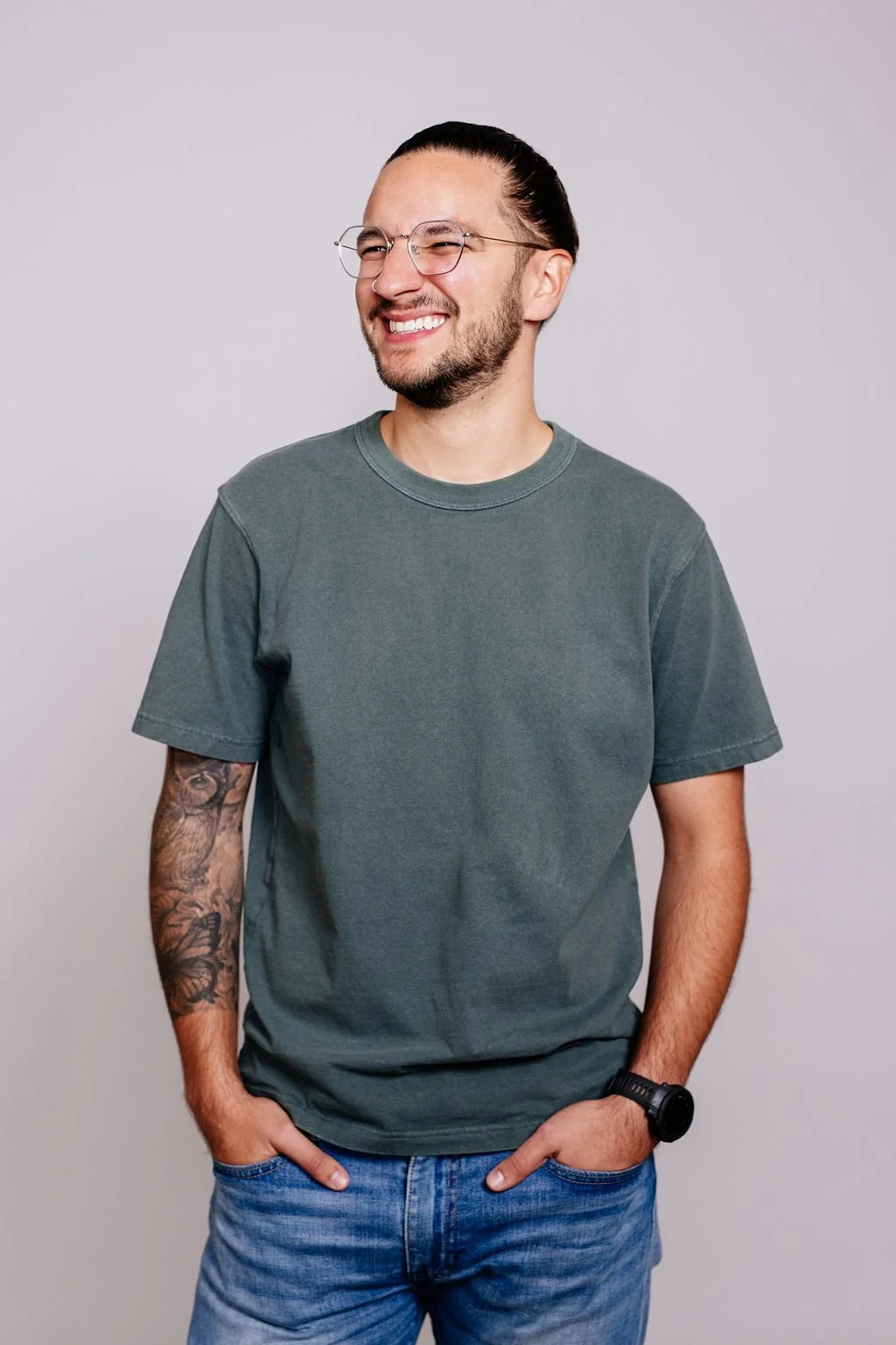 Smiling young man with glasses, tattooed arm, wearing a green t-shirt, blue jeans, and a watch, standing against a plain gray background.