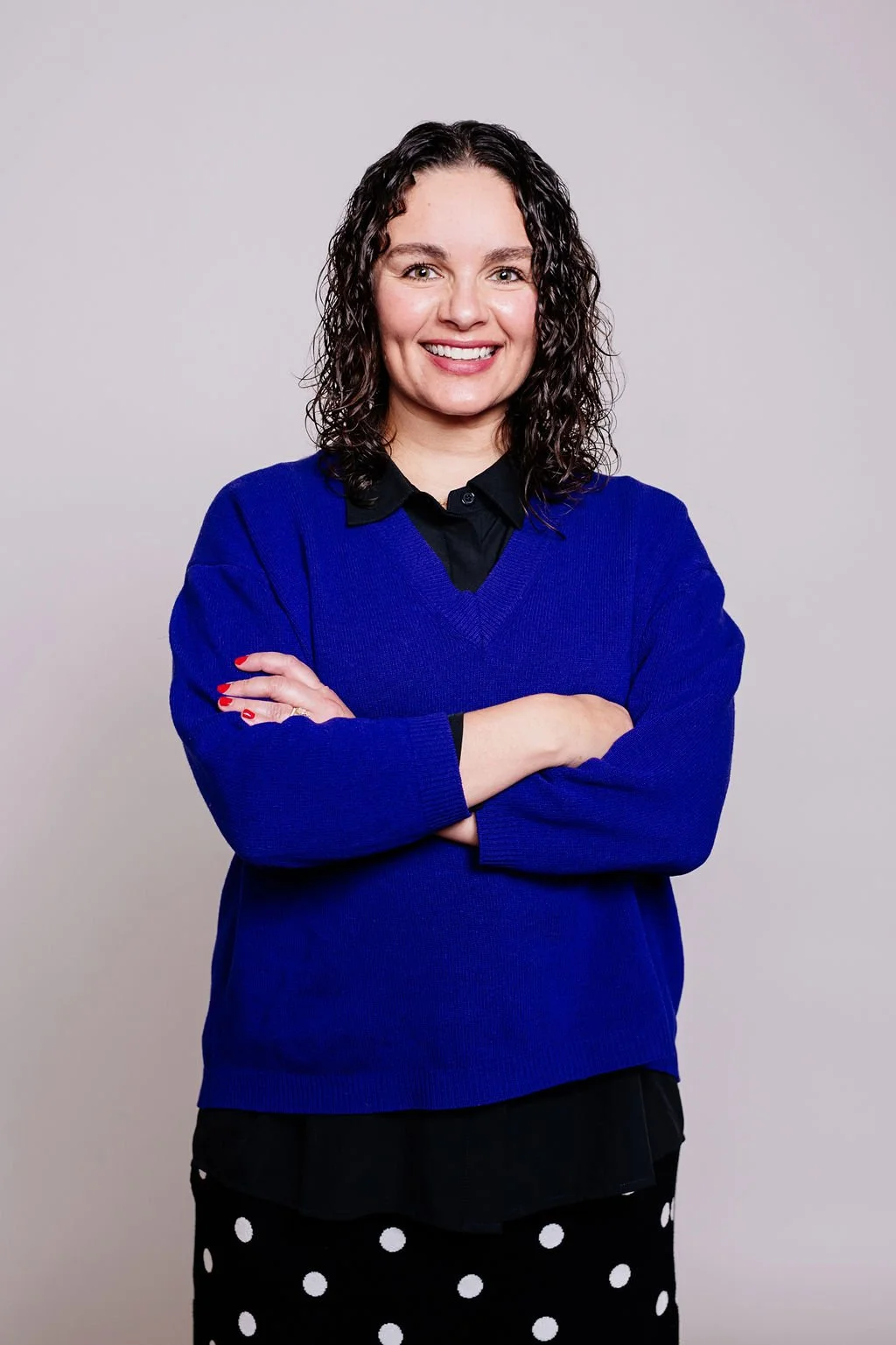 A woman with curly dark hair smiling and crossing her arms, wearing a blue sweater over a black collared shirt, and black pants with white polka dots, standing against a plain light gray background.