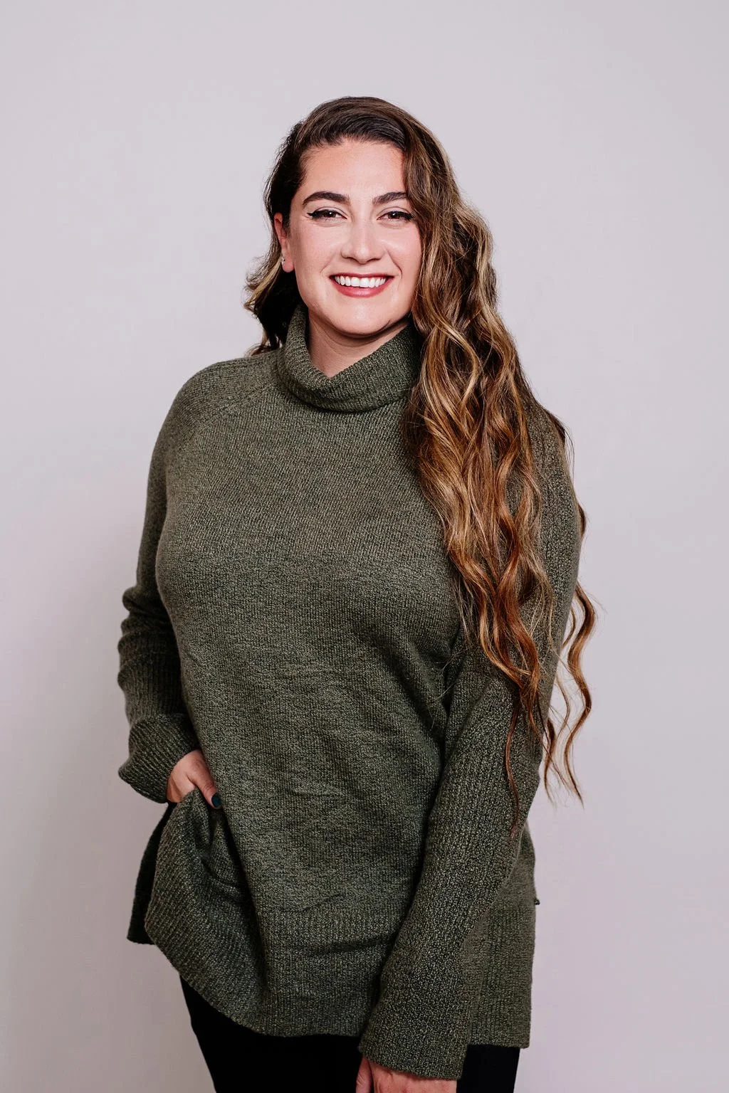 A woman with long, wavy light brown hair, smiling while wearing a green turtleneck sweater against a plain light gray background.