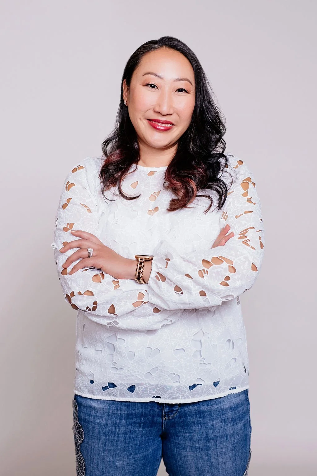 A woman with long black and red hair, smiling, wearing a white lace long-sleeved top and blue jeans, standing against a plain light gray background.