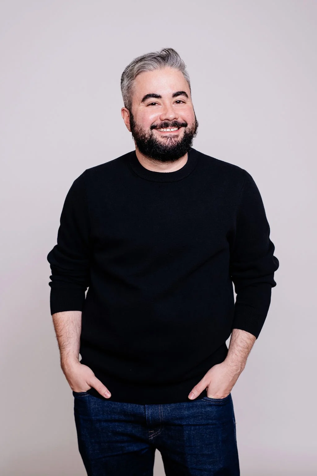 A smiling man with gray hair and a beard wearing a black sweater and blue jeans against a plain light gray wall.