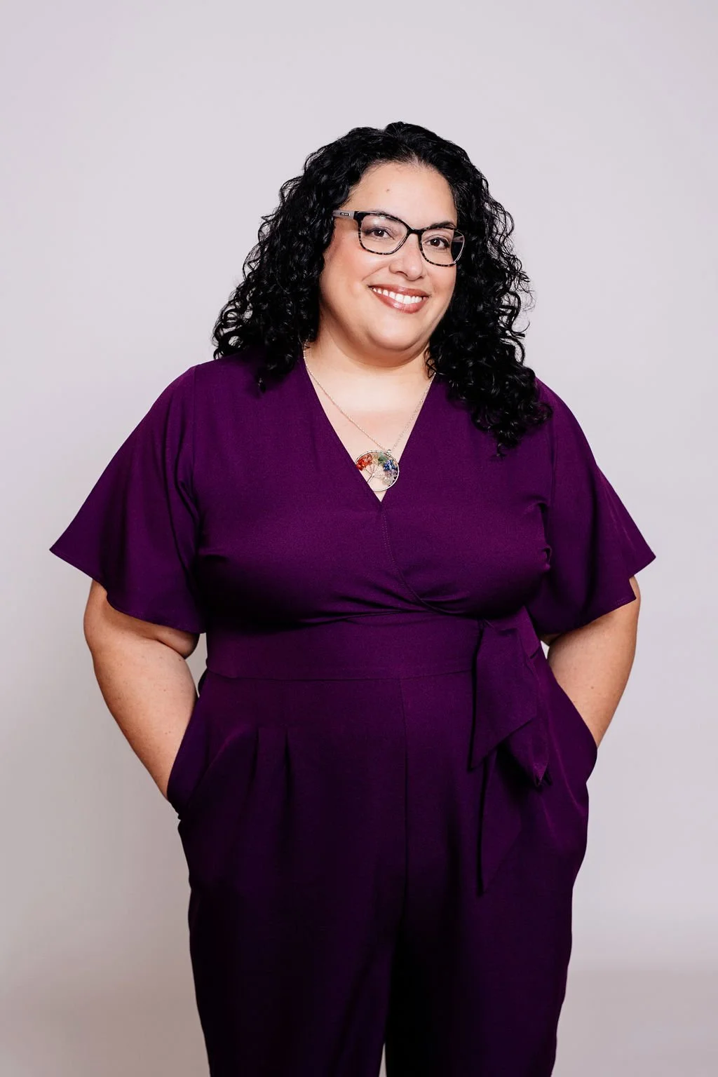 A woman with black curly hair, wearing glasses and a purple dress, smiling with her hands in her pockets against a light gray background.