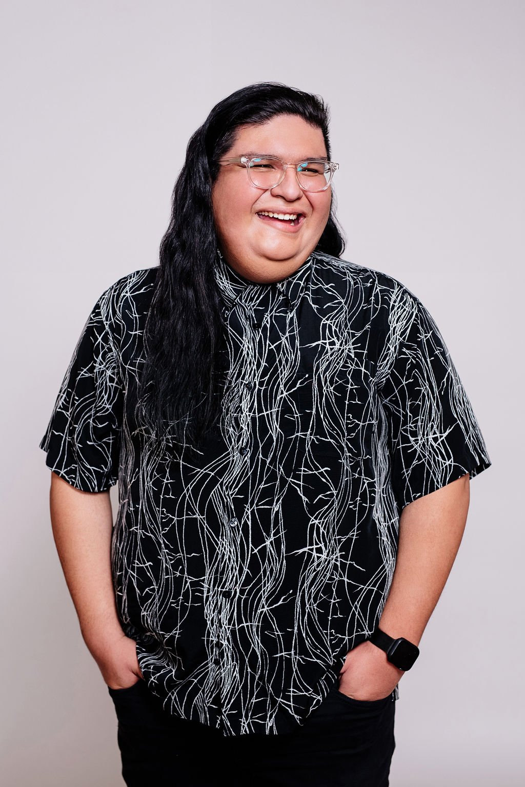 A person with long black hair, glasses, and a patterned black and white shirt, smiling and standing with hands in pockets against a plain white background.