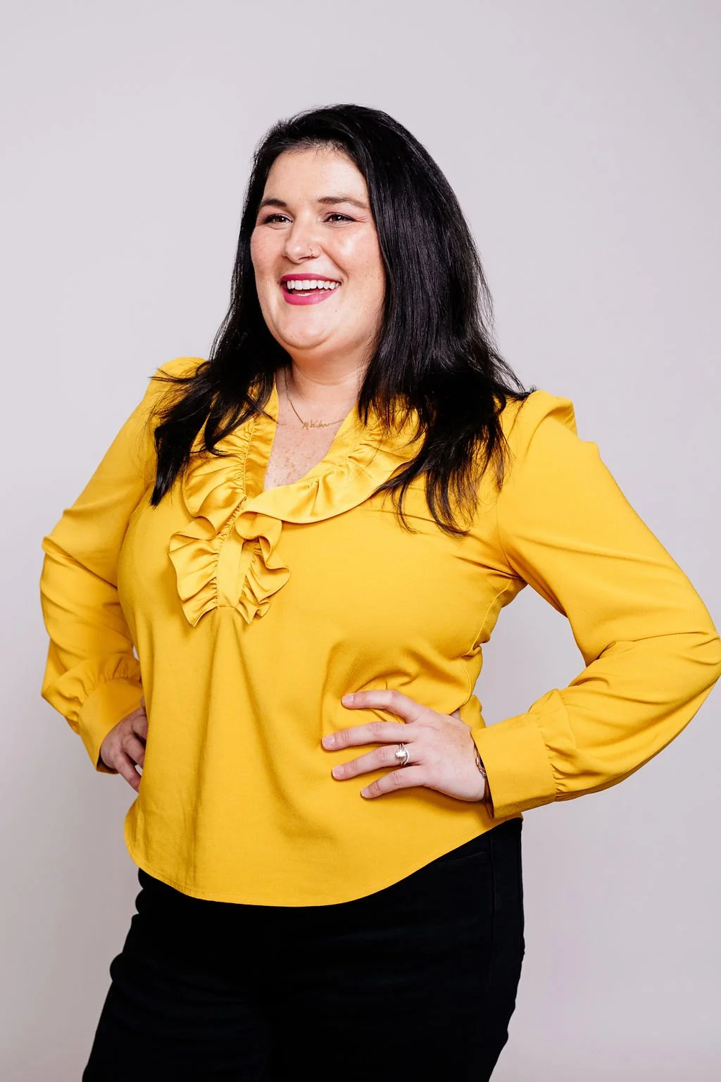 Woman with long black hair, wearing a yellow blouse with ruffled collar and black pants, smiling and standing against a plain light background.