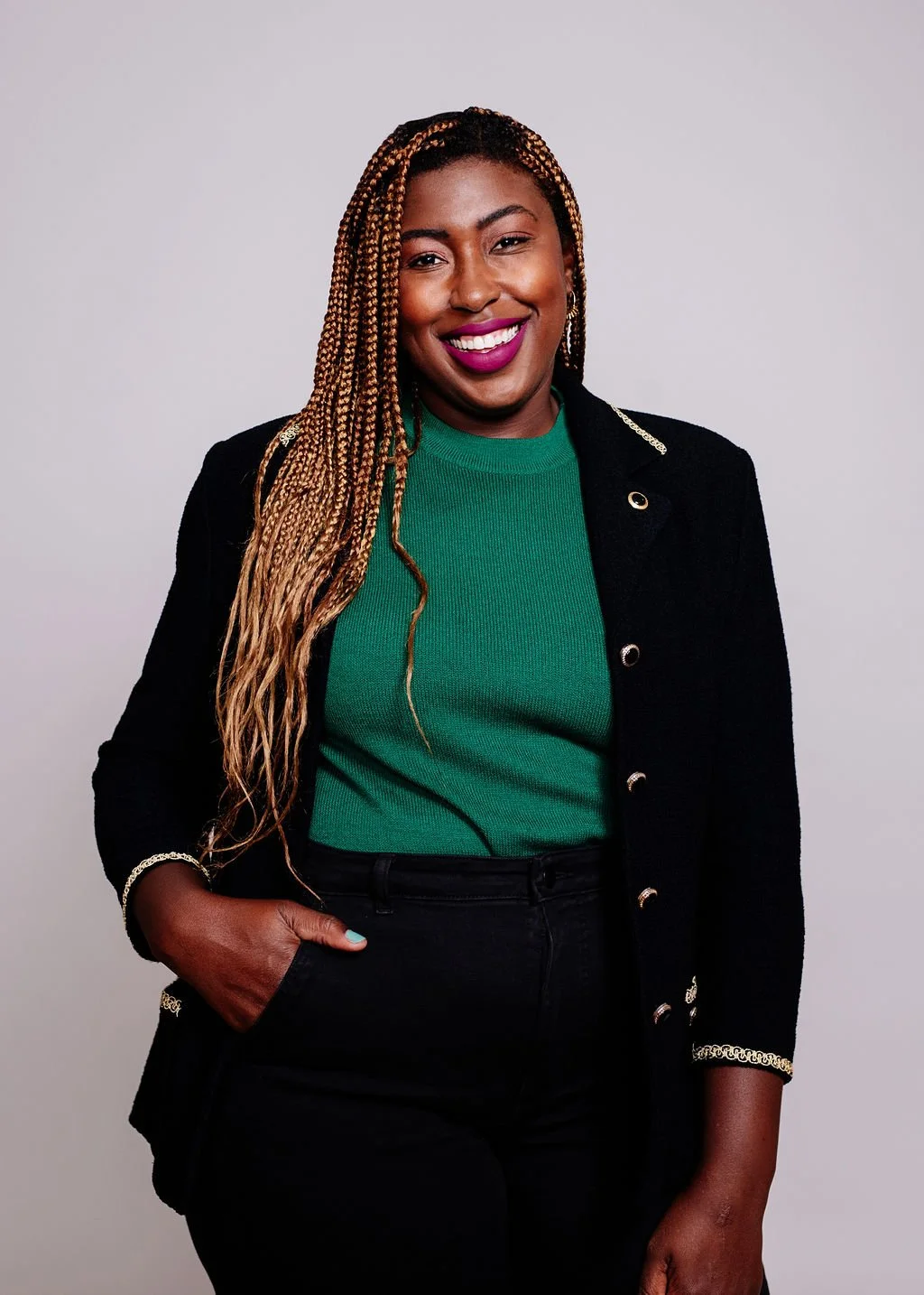 A woman with long blonde braids, smiling, wearing a green top, black blazer with gold detailing, and black pants, standing against a light gray background.