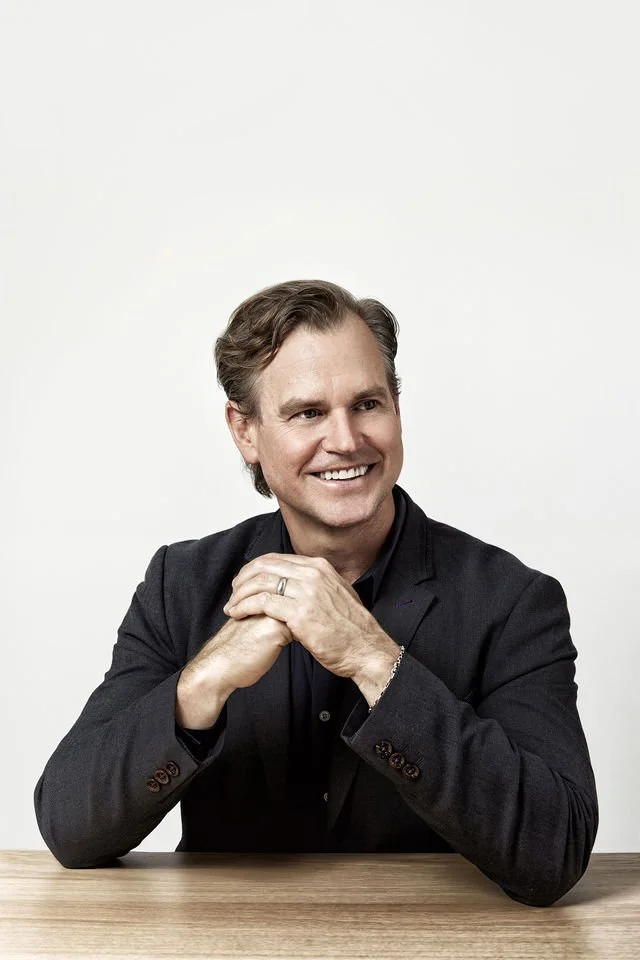 A smiling man in a dark blazer sits at a wooden table with hands clasped, against a plain white background.
