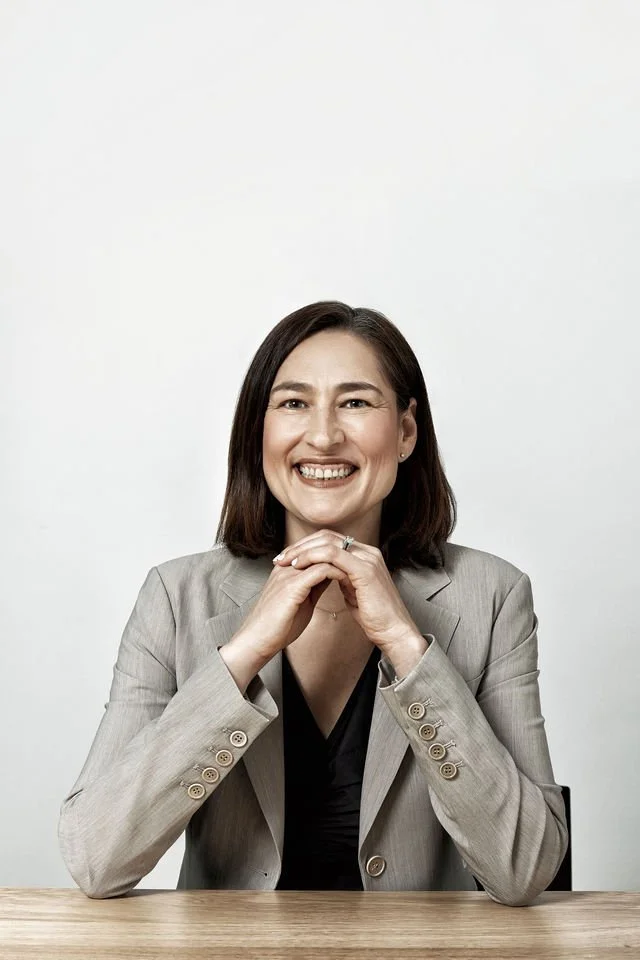 A smiling woman with shoulder-length brown hair, wearing a light gray blazer and dark blouse, sitting at a wooden table against a plain white background.