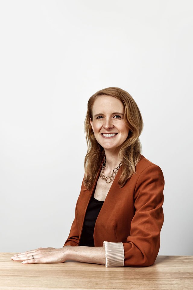 Smiling woman with shoulder-length red hair, wearing a rust-colored blazer and a chunky necklace, sitting at a table against a plain white wall.
