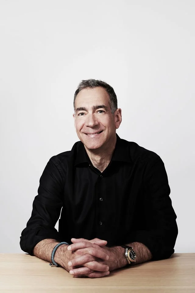 A smiling middle-aged man with dark hair, sitting at a table, wearing a black button-up shirt and a watch, against a plain light-colored background.