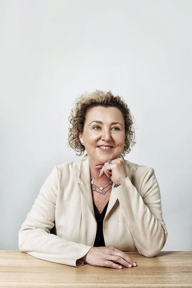 A woman with curly blonde hair sitting at a wooden table, smiling, with her chin resting on her hand, wearing a light-colored blazer and jewelry, against a plain white background.