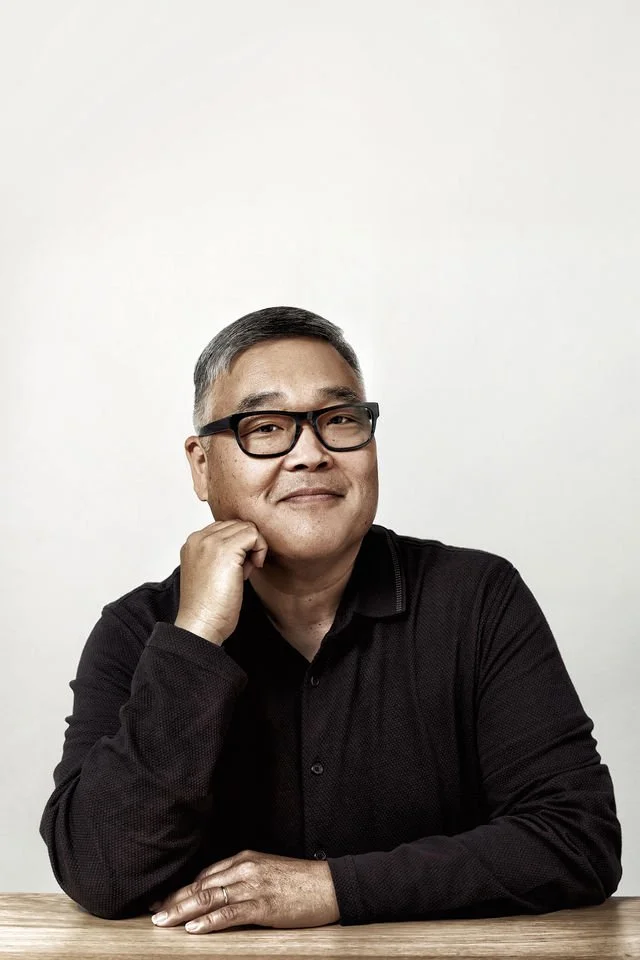 A middle-aged man with short gray hair and black glasses, wearing a black long-sleeve shirt, sitting at a wooden table with his chin resting on his hand and smiling at the camera, against a plain white background.