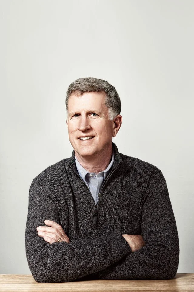A middle-aged man with short gray hair, smiling, wearing a dark gray zip-up sweater over a light blue collared shirt, sitting with arms crossed in front of a plain light background.
