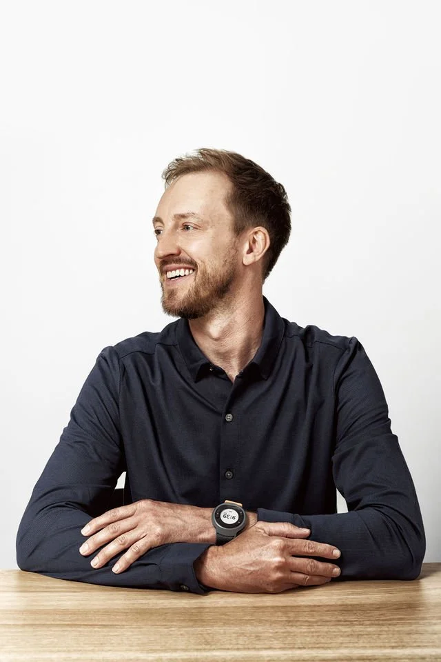 A smiling man with a beard, wearing a dark long-sleeve shirt and a watch, sitting at a wooden table against a plain white background.